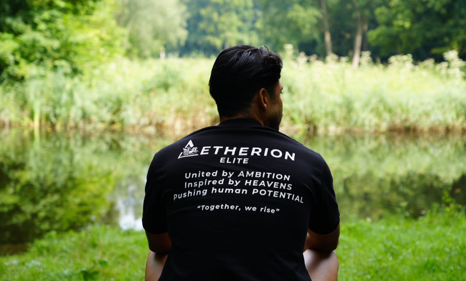 Person wearing an 'Aetherion Elite' t-shirt by a lake with greenery.